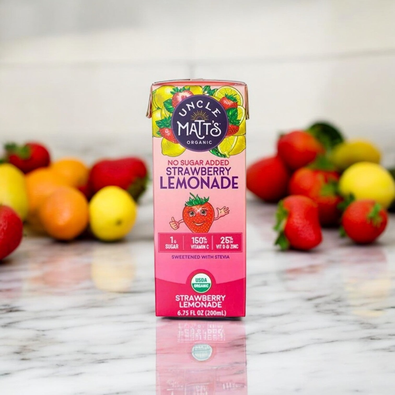 A carton of Uncle Matt's Strawberry Lemonade Juice Box - No Sugar Added, 6.75 oz, sits on a marble surface, with strawberries and lemons in the background. Packed with essential vitamin C, this refreshing drink is perfect for any occasion.