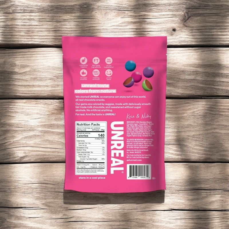 A pink bag of Unreal Candy Coated Milk Chocolate Gems, 6 oz - 1 Count by Unreal is placed on a wooden surface. The front of the bag features colorful candies, a nutritional facts label, and text describing the product made with organic ingredients for a guilt-free indulgence.