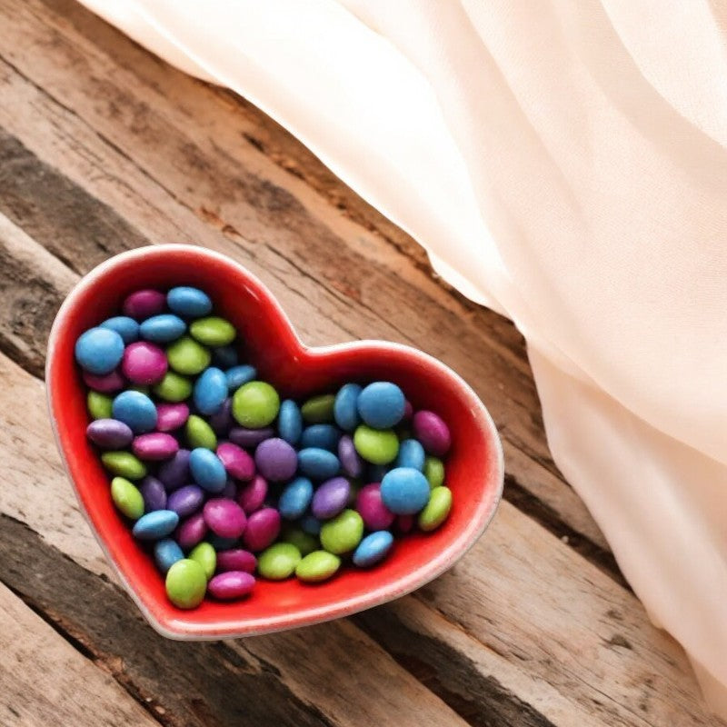 An Unreal heart-shaped bowl filled with Unreal Candy Coated Milk Chocolate Gems, 6 oz - 1 Count, sits on a rustic wooden surface with a white cloth draped nearby.