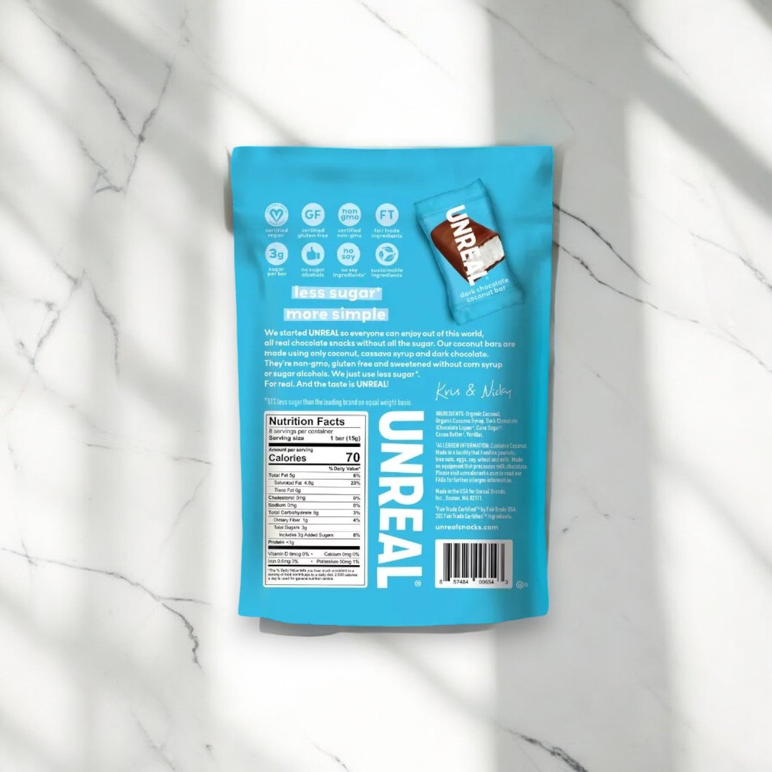 A blue package of Unreal Dark Chocolate Coconut Bars - 1 Count is displayed on a marble surface, highlighting nutritional information and certifications such as non-GMO, vegan, gluten-free, and fair trade. The bag features an image of a single candy piece.