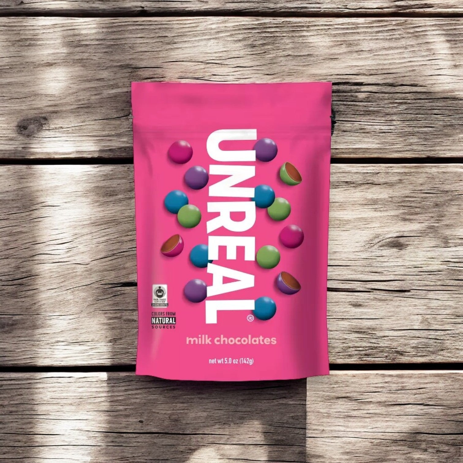 A pink pouch of Unreal Candy Coated Milk Chocolate Gems, 6 oz - 1 Count, featuring colorful candy-coated chocolates on its exterior, made with fair trade chocolate and organic ingredients by Unreal, placed on a wooden surface.