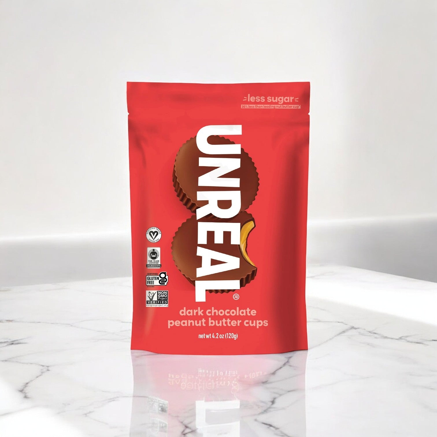 A bag of Unreal Dark Chocolate Peanut Butter Cups, 4.2 oz - 1 count, is displayed on a marble surface. The red packaging prominently displays product information and logos indicating that it is non-GMO, fair trade certified, vegan, and gluten-free.