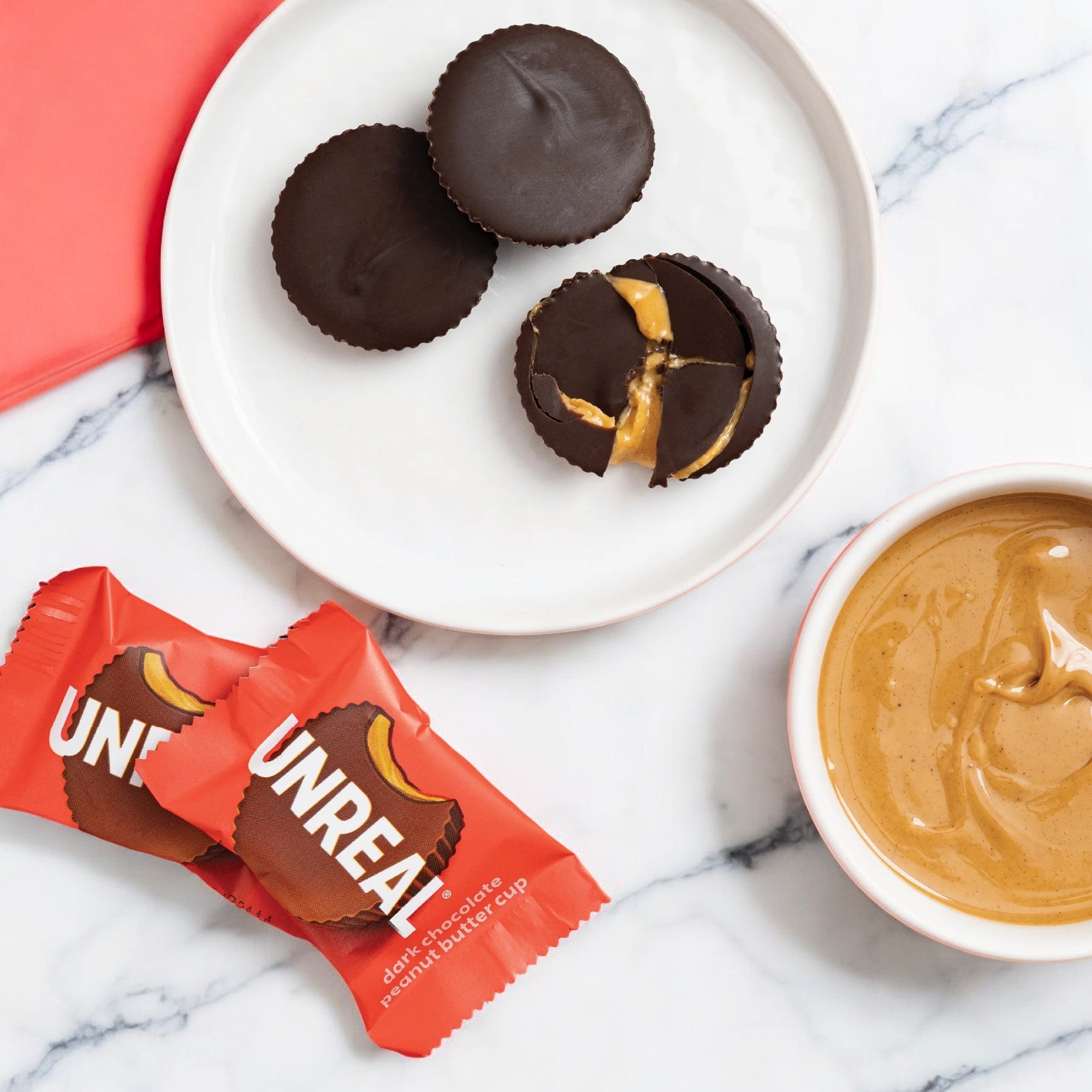 Two unwrapped Unreal Dark Chocolate Peanut Butter Cups sit on a plate, with one broken to reveal the creamy peanut butter filling inside. Beside a bowl of peanut butter on a marble surface are two wrapped cups. These Unreal vegan delights are also gluten-free, making them perfect for any indulgent yet mindful snacking moment.