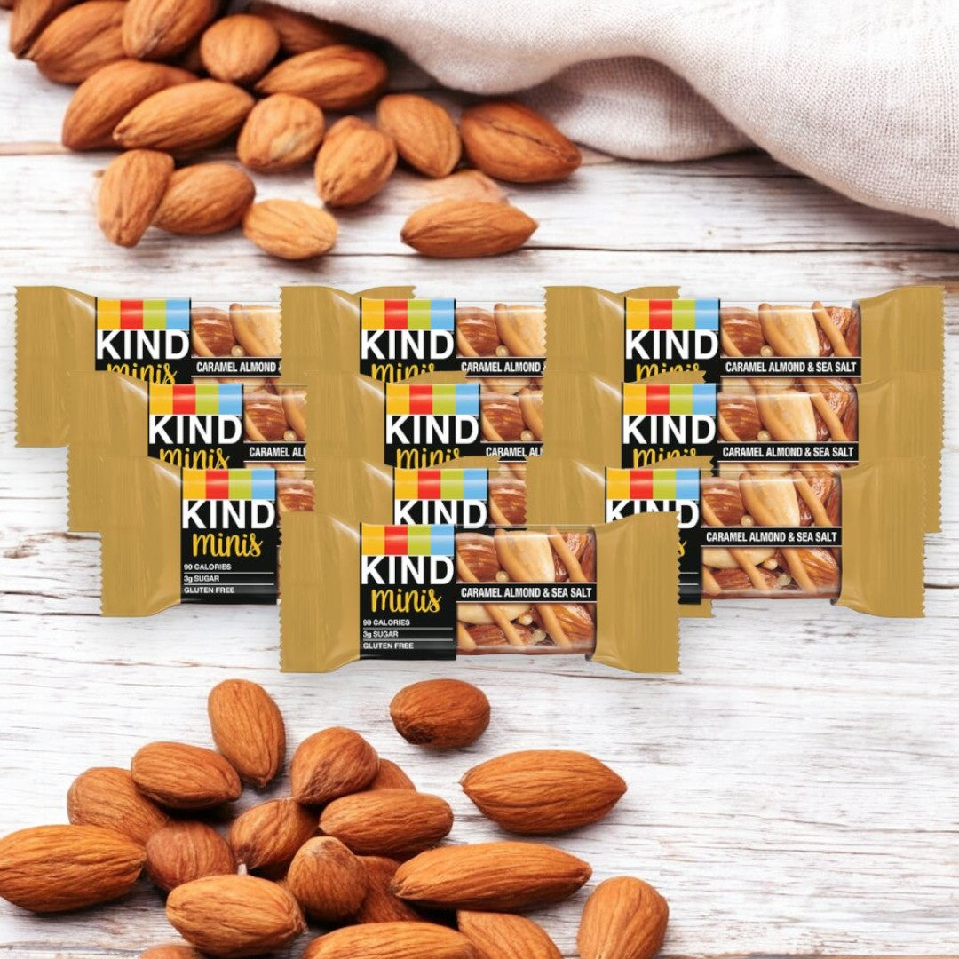 Ten Kind Minis Caramel Almond & Sea Salt bars are displayed with almonds scattered around on a wooden surface. In the background, a cloth bag spills out almonds, showcasing this healthy snack option from the Kind brand.