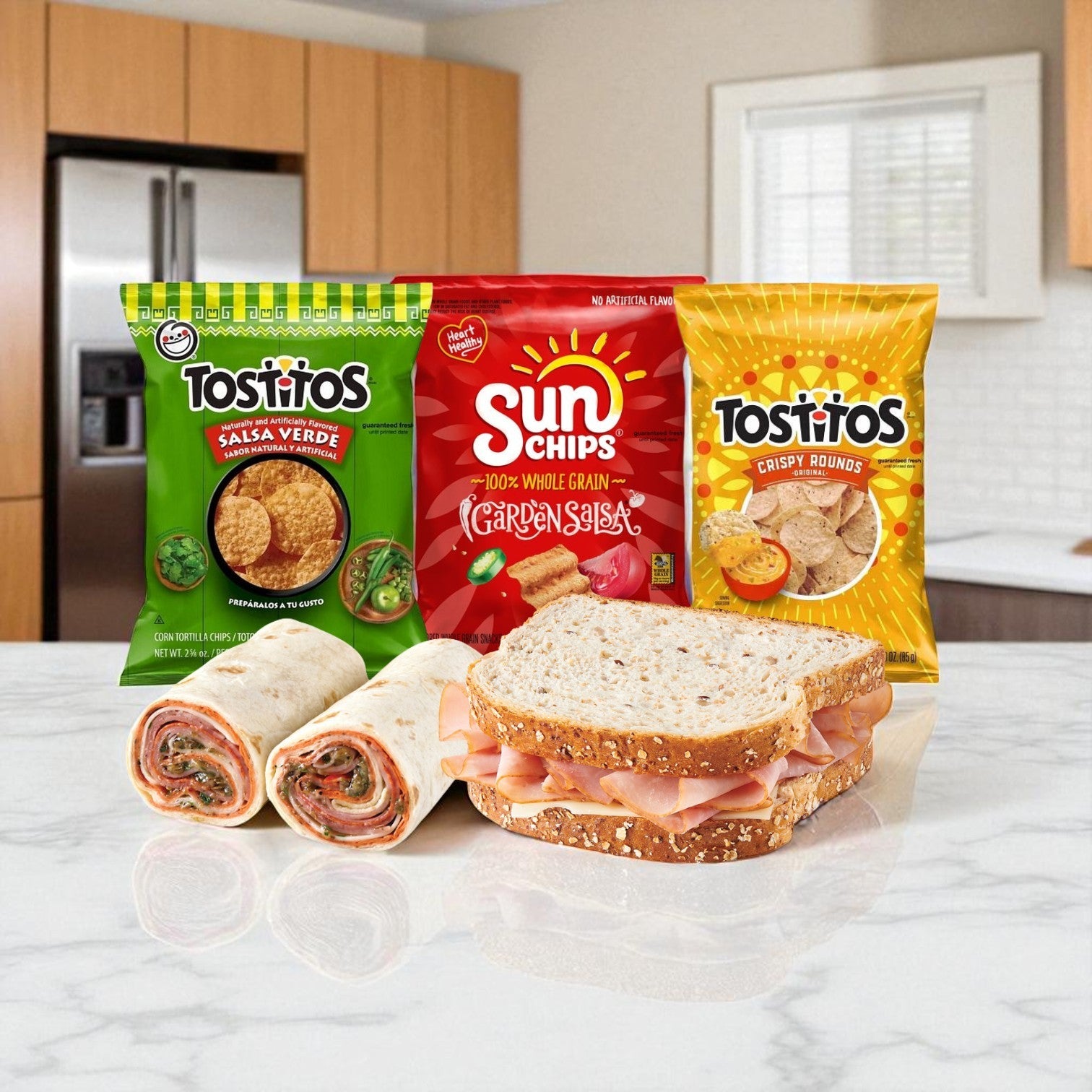A Snack & Deli Variety Pack by E.A. Sween, featuring three bags of chips, a deli meat sandwich on whole wheat, and two ready-to-eat wraps, is displayed on a kitchen counter with a blurred kitchen in the background.