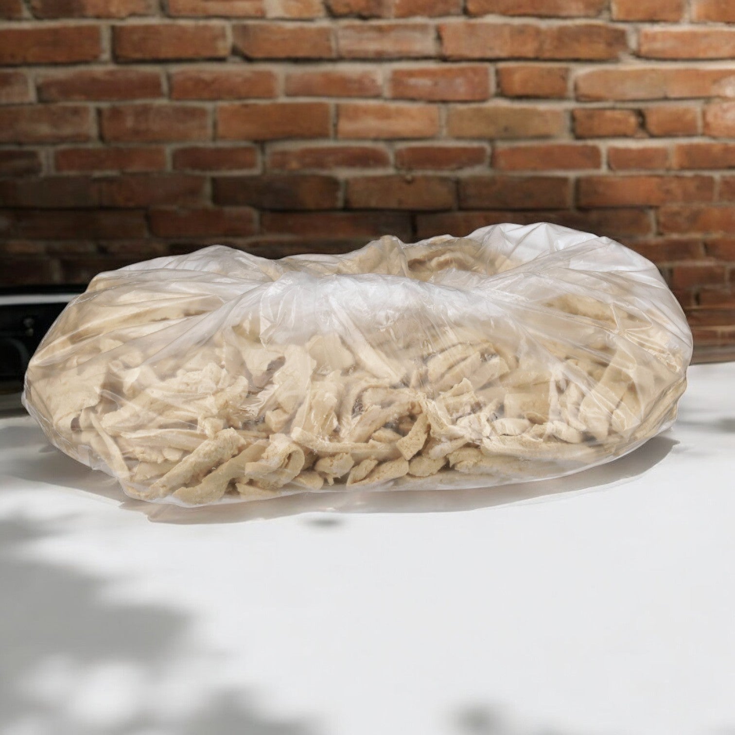 A large, clear plastic bag containing light-colored Gardein Veggie Burger Vegetarian Vegetable Chicken Strips from Beyond Meat rests on a white surface, set against a brick wall in the background.