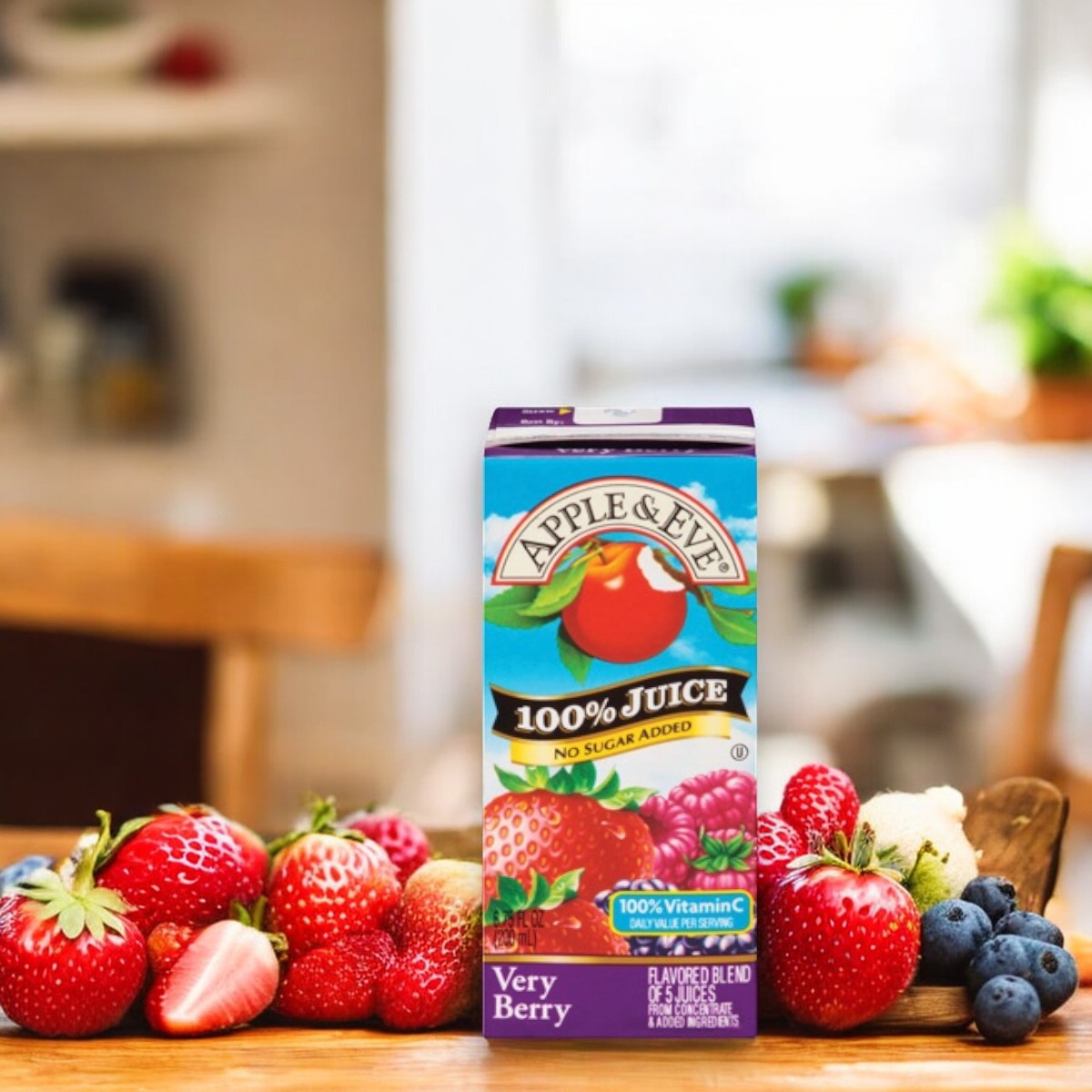 A carton of Adam and Eve Apple Juice, Very Berry Flavor, enriched with natural ingredients, is placed on a table surrounded by fresh berries such as strawberries, raspberries, and blueberries. The background showcases a blurred kitchen scene, emphasizing this healthy beverage from the Apple & Eve brand.