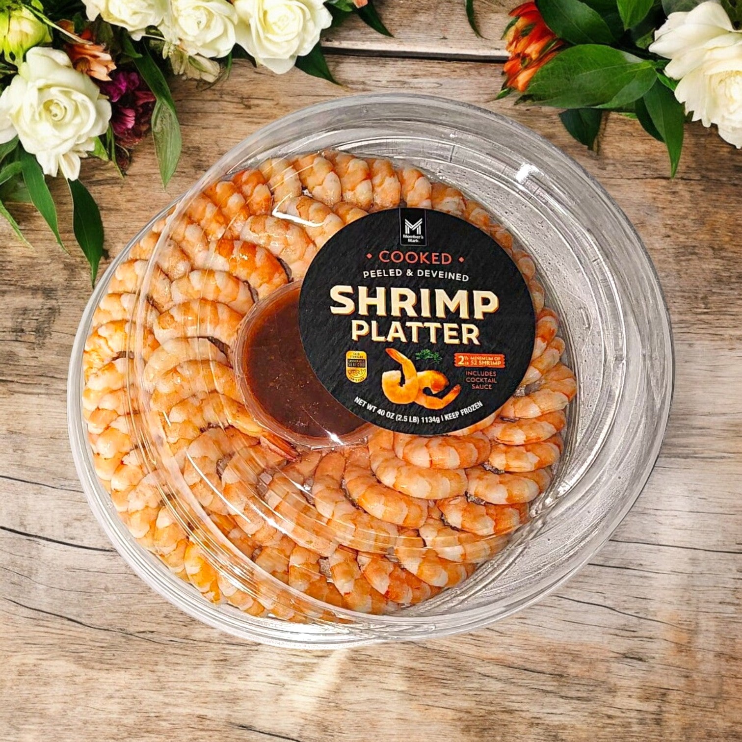 The Member's Mark Shrimp Platter with Cocktail Sauce, 2.5 lbs per pack (total 7.5 lbs), is elegantly presented in a round clear container on a floral-decorated wooden background. Enjoy premium seafood with free shipping!.