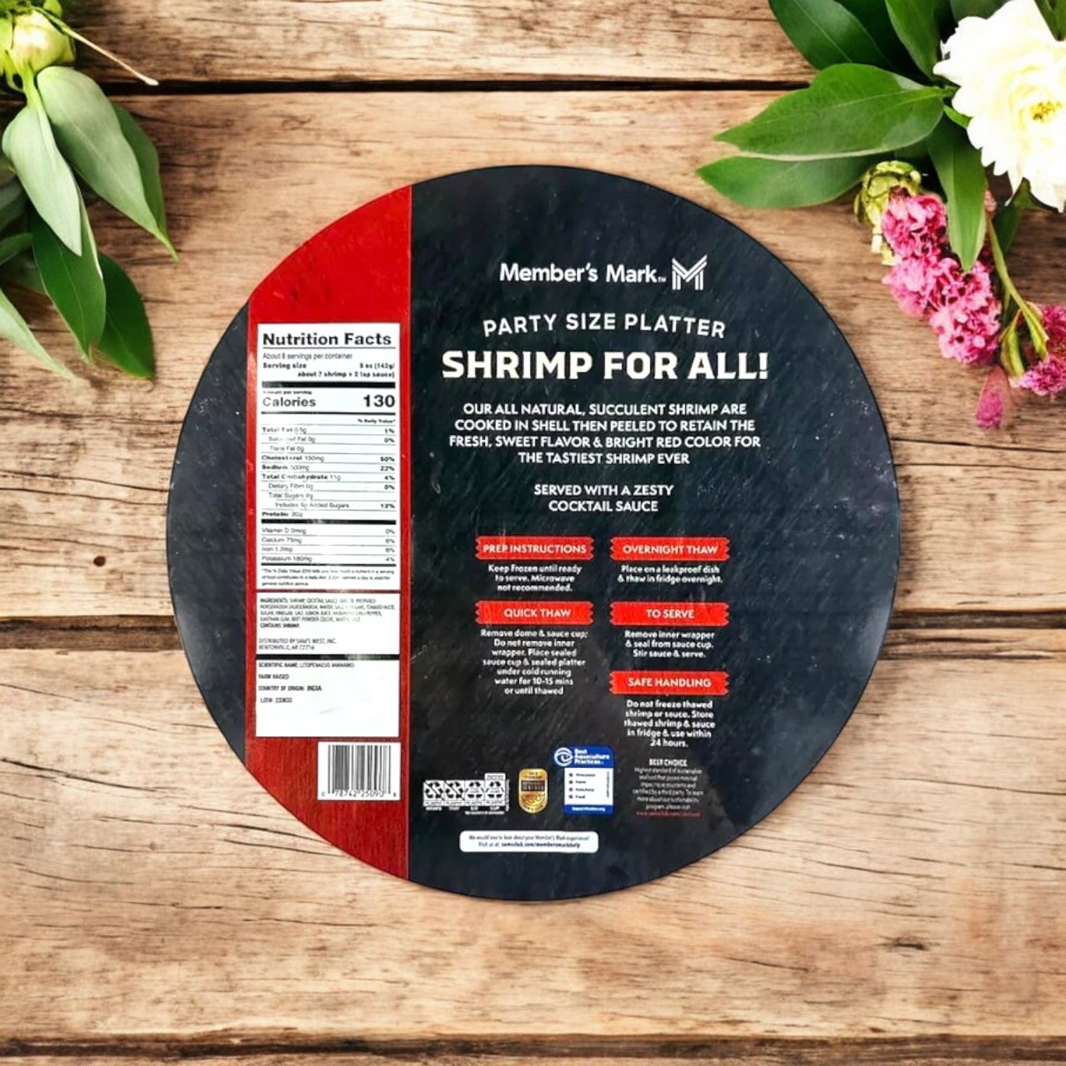 A round platter labeled "Shrimp for All!" sits amid flowers on a wooden table, with nutrition facts and preparation instructions. The Member's Mark Shrimp Platter with Cocktail Sauce, Frozen – 2.5 lbs, is an ideal premium seafood delight when paired with cocktail sauce for extra flavor.