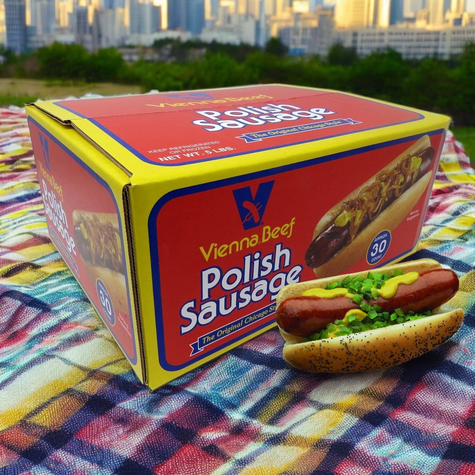 A 5-pound box of Vienna Beef Polish Sausage rests on a colorful cloth, with a hot dog topped with onions and chives in the foreground; the cityscape provides a perfect backdrop to this display of authentic flavor.