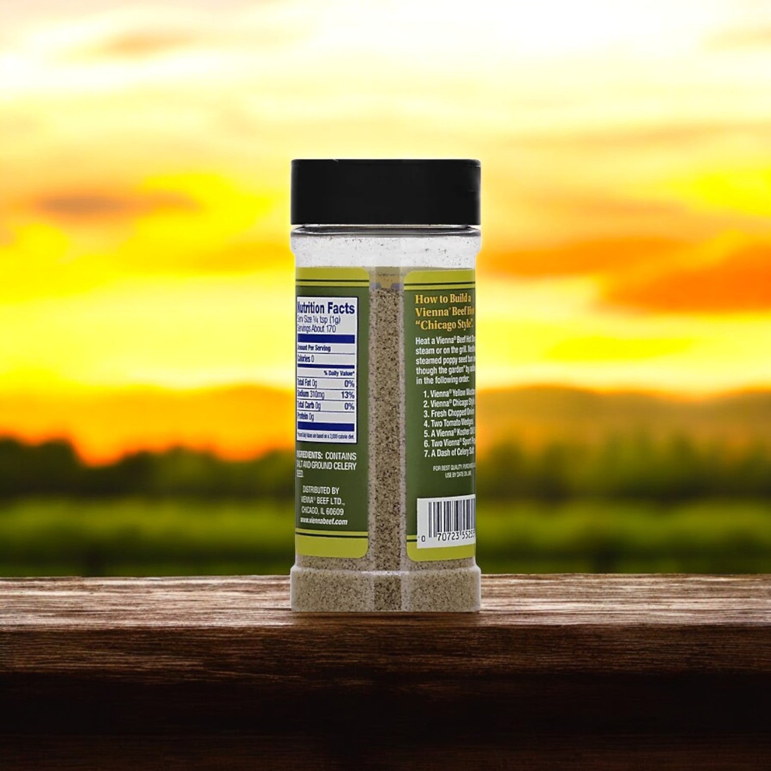 A Vienna Celery Salt 6 oz. bottle with labels and a black cap rests on a wooden surface against a blurred sunset backdrop. This Vienna Beef product is perfect for flavoring dishes, such as Chicago-style hot dogs, with limitless potential.