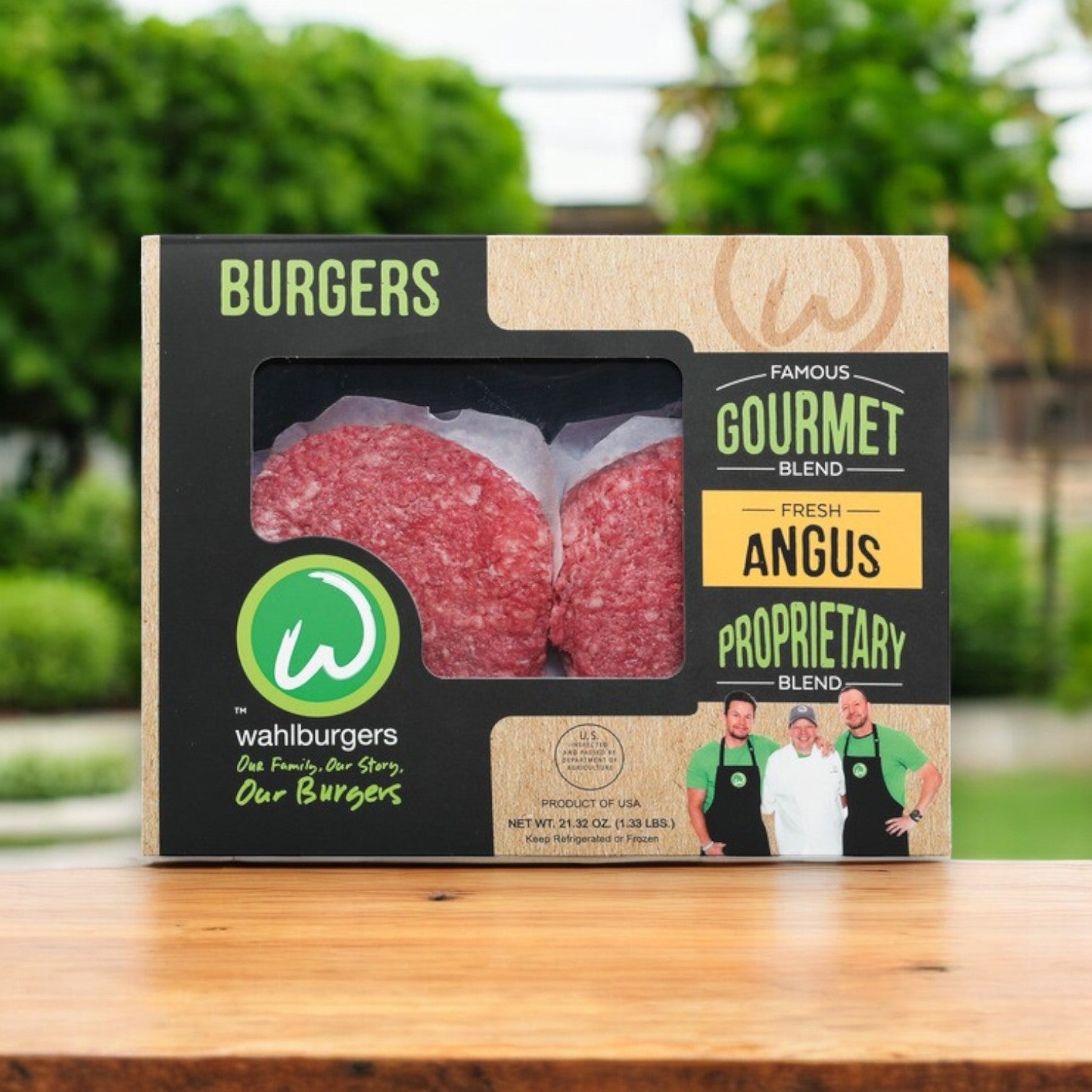 A box of Wahlburgers Famous Gourmet Blend Fresh Angus Burgers, 21.32 oz, sits on a wooden table with trees in the background. The packaging showcases the "Gourmet Blend" label and features images of three people in green shirts.