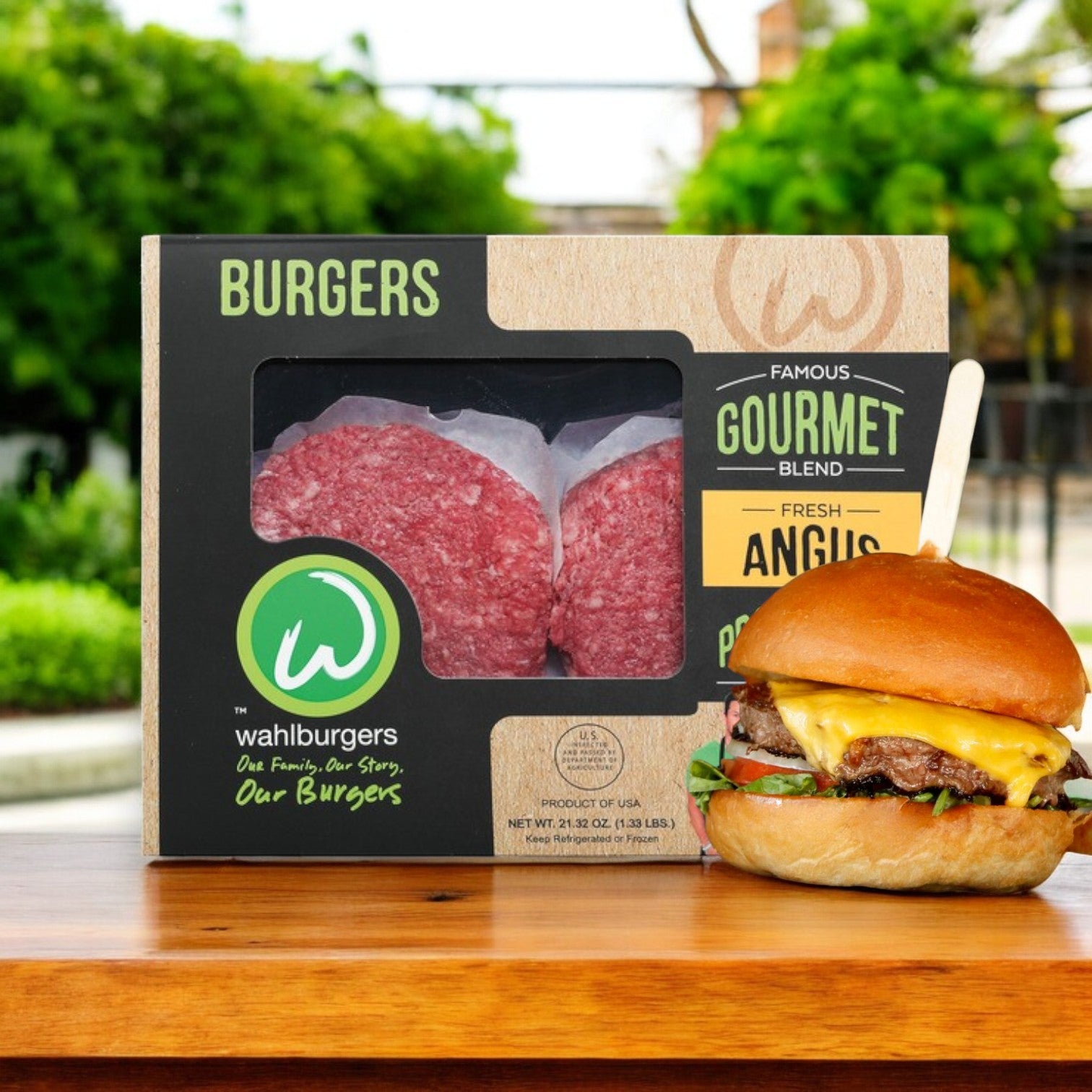 A box of Wahlburgers Famous Gourmet Blend Fresh Angus Burgers, 21.32 oz, sits next to a freshly made Angus cheeseburger with lettuce and tomato on a bun, placed on a wooden surface outdoors and surrounded by lush greenery.