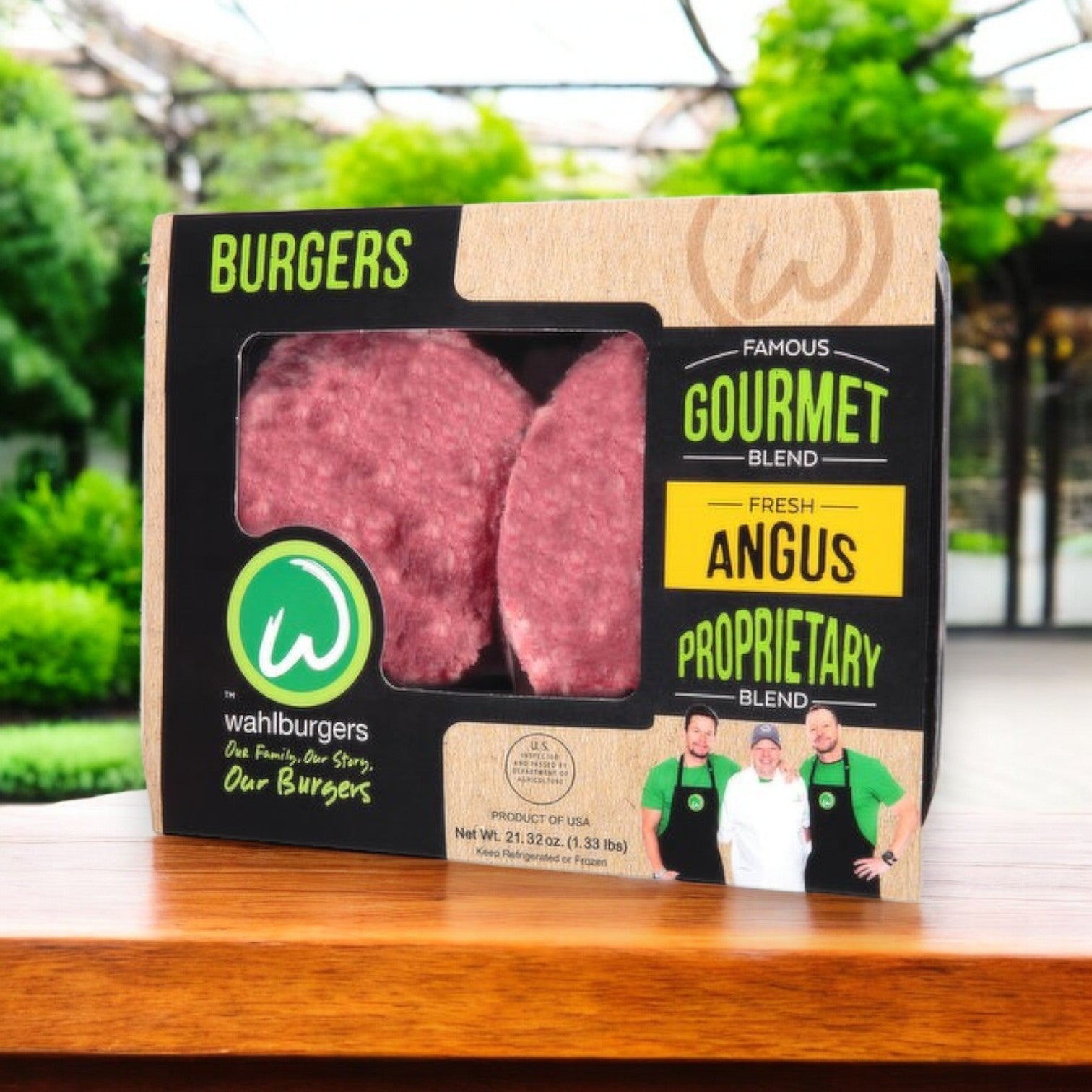 The packaging of Wahlburgers Famous Gourmet Blend Fresh Angus Burgers, 21.32 oz, rests on a wooden table featuring images of dedicated staff and labels promoting their fresh Angus burgers, set against a vibrant backdrop of lush outdoor greenery.