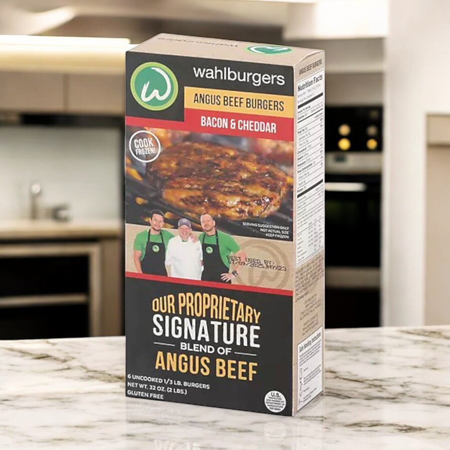 A box of Wahlburgers Frozen Beef Patties, Bacon & Cheddar, 32 oz rests on the kitchen counter, highlighting a tempting image of cooked Angus beef burgers with bacon and cheddar alongside three people in uniforms.