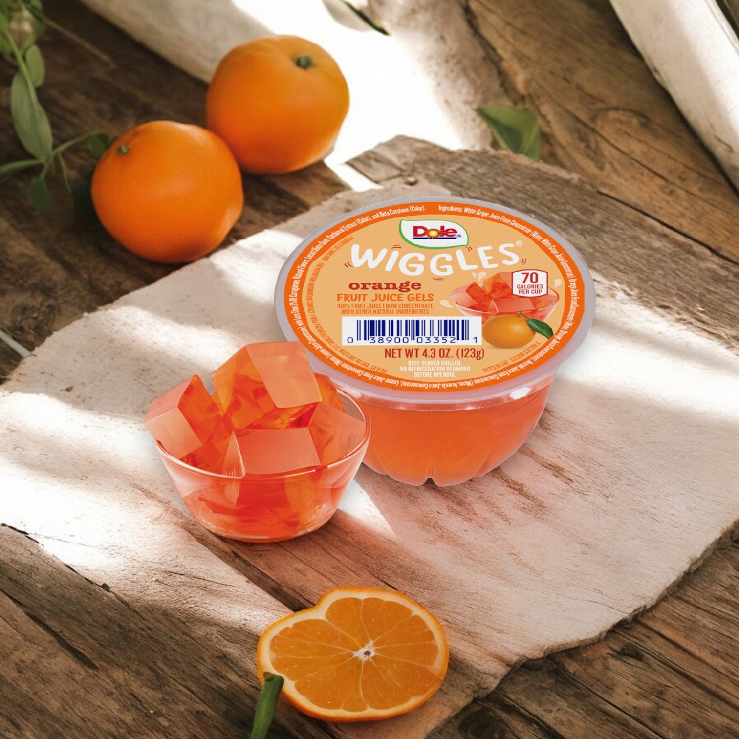 A single 4.3 oz container of Dole Wiggles Orange Fruit Juice Gels, known for its no added sugar and high Vitamin C content, is placed on a wooden surface next to an open cup displaying the cubed gels, with two oranges in the backdrop.