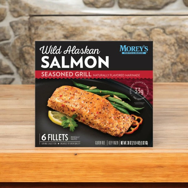 Box of Morey's Fine Fish & Seafood's Marinated Wild Alaskan Salmon, 6 ct - 1 Box, placed on a wooden surface with a stone backdrop. The packaging showcases the six marinated salmon fillets along with nutrition information and a gluten-free label. Discover the taste of premium seafood with every bite.