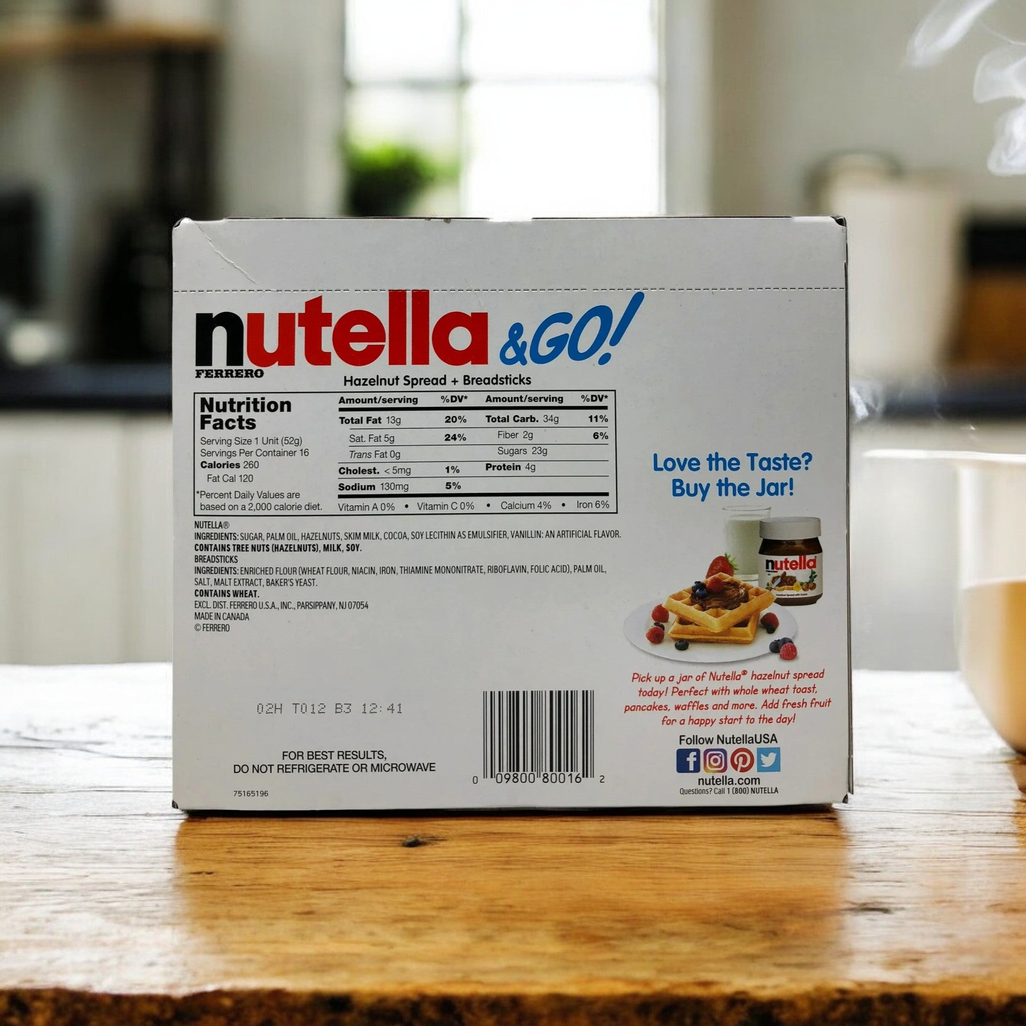 The Nutella and Go Hazelnut Spread with Breadsticks Ferrero 16 Pack - 1.8 oz Each from Easy Lunches is placed on a table, displaying the nutritional facts and product information on the back. In the blurred background, there is a cup of coffee and a plant.