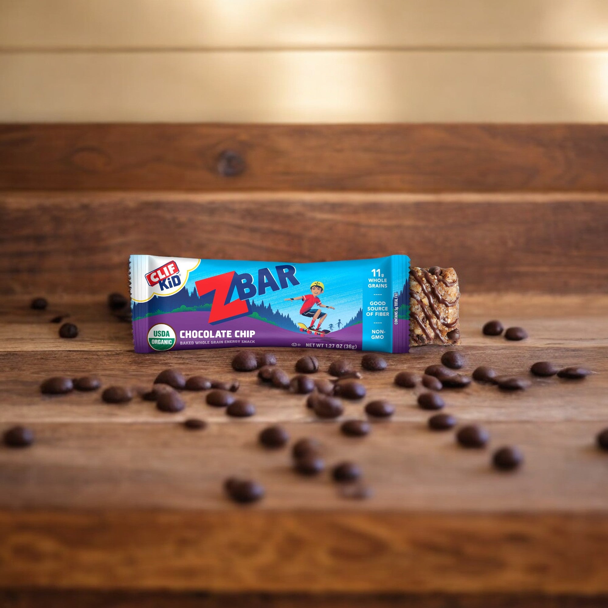 A Clif Bar Z Bar Chocolate Chip from Clif Kid, perfect for on-the-go snacking, is placed on a wooden surface with scattered chocolate chips around it. The chewy snack bar’s wrapper is blue and purple with a design of a child skateboarding, highlighting its wholesome whole grains.