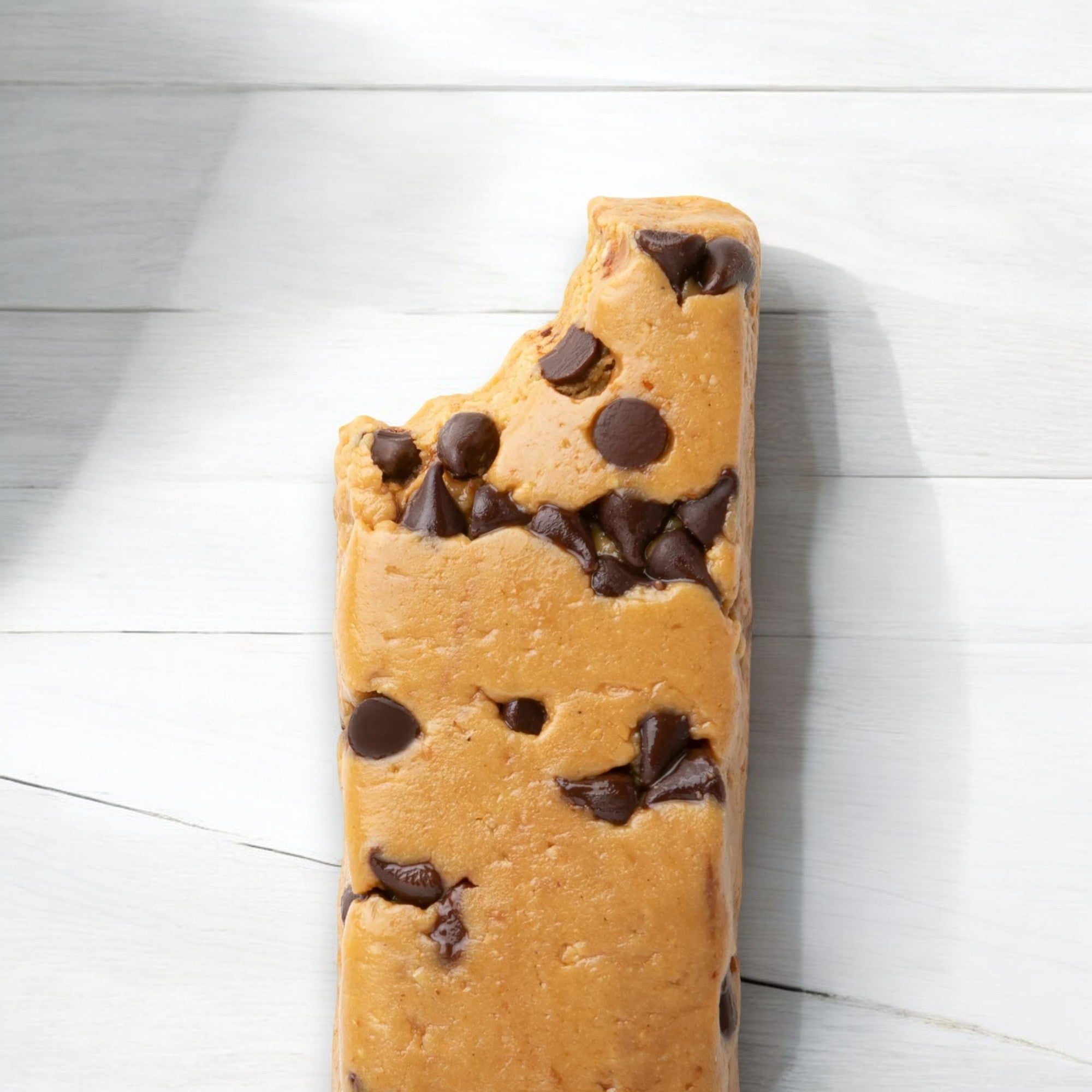 A Perfect Bar Gluten-Free Dark Chocolate Chip Peanut Butter Protein Bar, 2.3 oz - 1 Count with a bite taken out, featuring dark chocolate chips and placed on a white wooden surface.