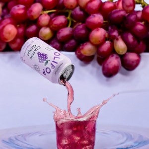 A can labeled "OLIPOP Classic Grape - 12 fl oz - 1 Count" from Olipop pours a pinkish liquid into a glass, causing a splash. Rich in prebiotics for digestive health, the beverage is complemented by a bunch of grapes visible in the background.