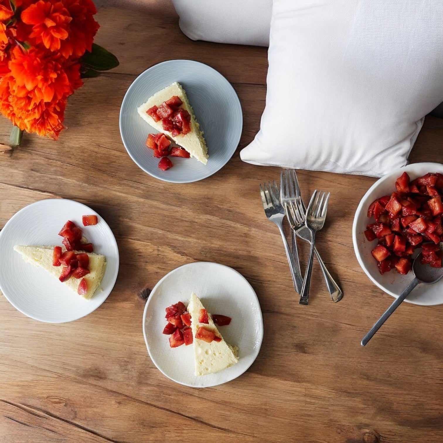 On a wooden table, there are three plates featuring Eli's Cheesecake Original Plain Single Serve, 3 oz., each topped with diced strawberries. The setup is completed with a bowl of additional strawberries, forks, and a bouquet of orange flowers.