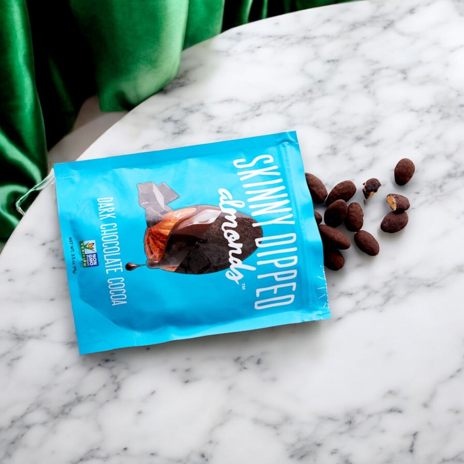 A blue bag of Skinny Dipped SkinnyDipped Dark Chocolate Candy Cocoa Almonds, labeled "Dark Chocolate Candy Cocoa Almonds Bag 3 oz," lies partly opened on a marble surface with some dark chocolate almonds spilled out, offering a tantalizing and healthy snack.