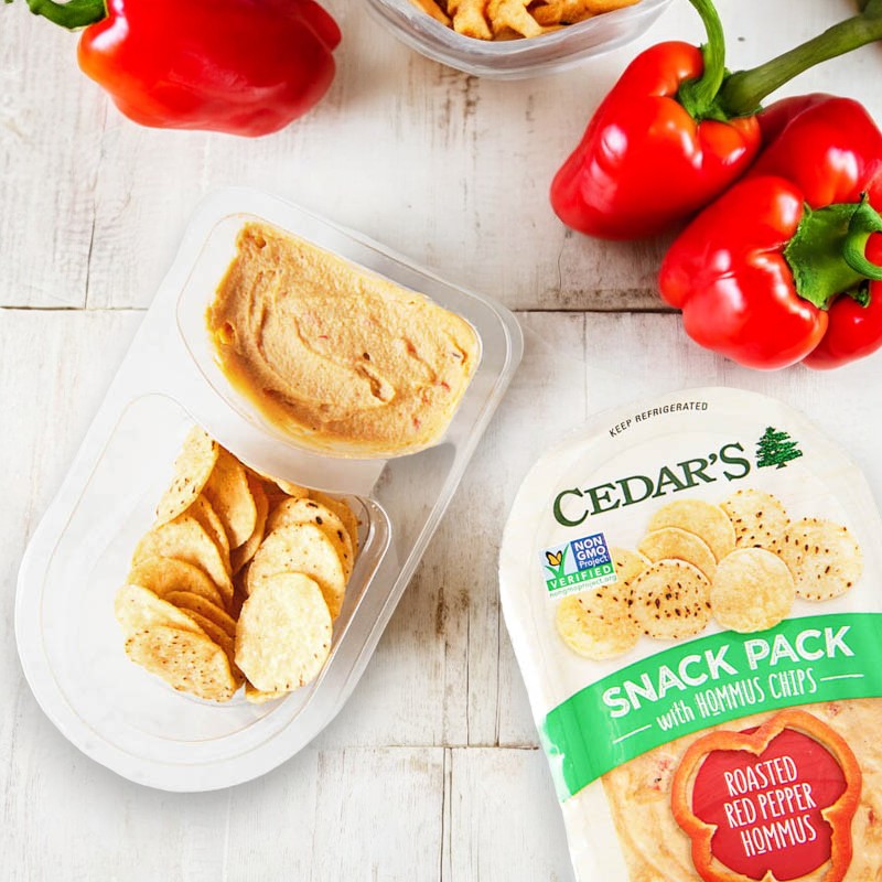 A snack pack of hummus with crispy chips is displayed on a wooden surface, accompanied by fresh red bell peppers. The label on the pack reads "Cedar's Roasted Red Peppers Hommus w/ Hommus Chips -1 Count.