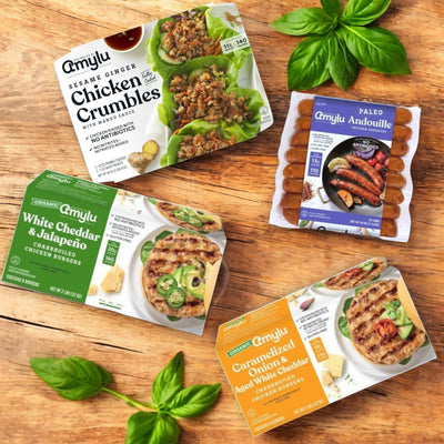 Four packaged foods rest on a wooden surface with basil leaves: Amylu’s 4-Pack Combo features Amylu Chicken Products like chicken crumbles, organic chicken burgers, white cheddar jalapeño options, and paleo-friendly andouille sausage caramelized onion burgers.