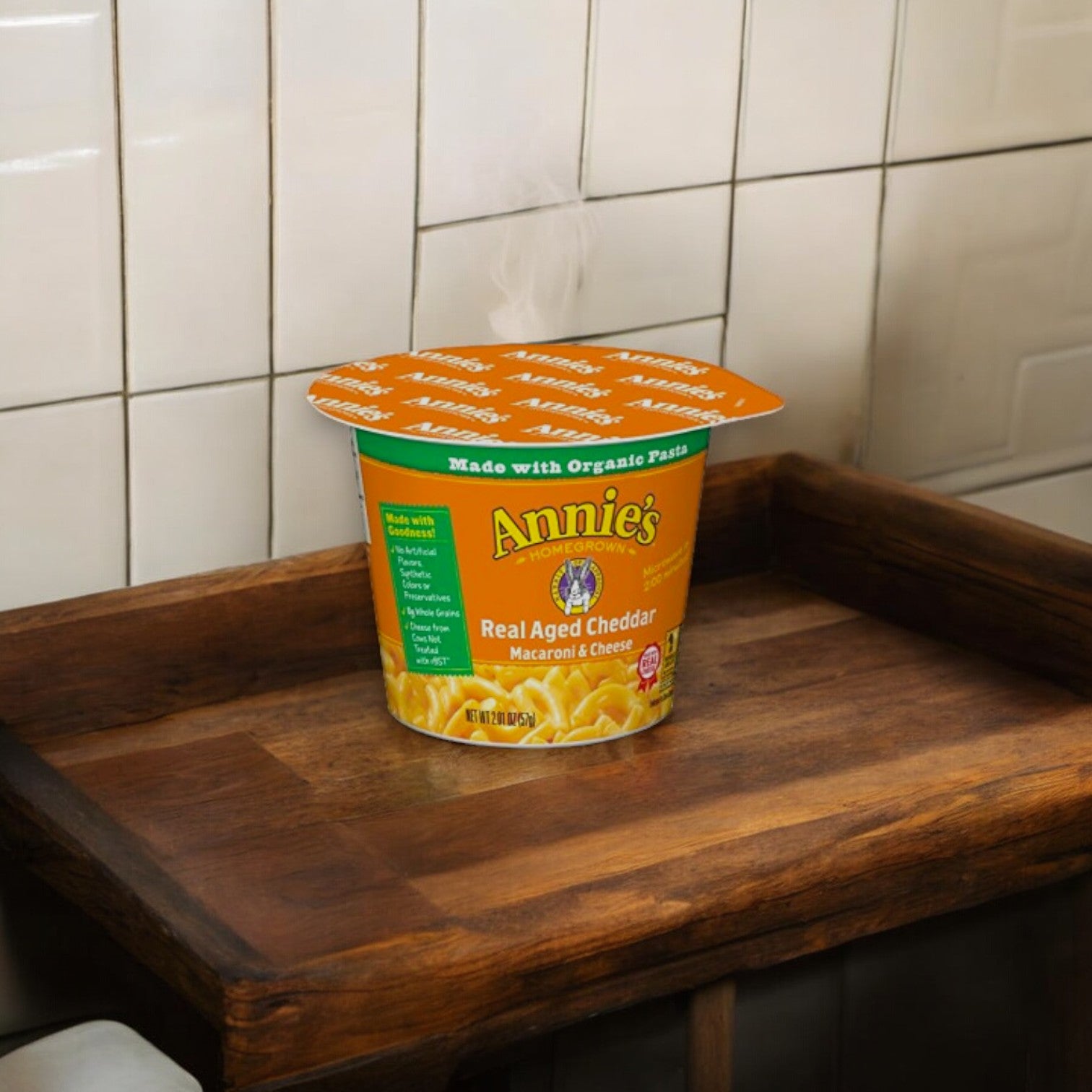 A steaming cup of Annie's Real Aged Cheddar Microwave Mac and Cheese, made with organic pasta, sits on a wooden tray against a tiled wall background.