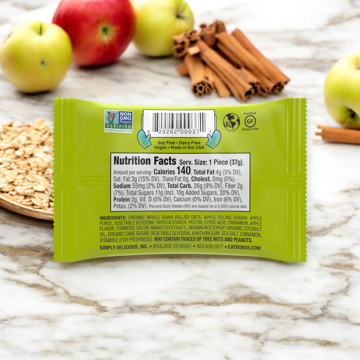 A package of Bobo's Stuff'd Apple Pie Bites (1.3 oz, 1 Count) with nutrition facts and ingredients list visible on the back. The package is surrounded by fresh apples, wholesome whole grain oats, and cinnamon sticks, all placed on a wooden surface.