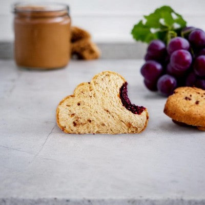 A slice of cloud-shaped bread with a bite taken out, revealing a purple filling. A bunch of grapes and a jar of spread are in the background, showcasing the wholesome ingredients of Chubby Snacks' Peanut Butter & Grape Jam Sandwich (2 oz - 1 Count) by Chubby.