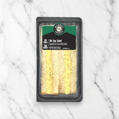 A plastic container with a green label and black details holds an E.A. Sween Dill Egg Salad On Homestyle White Bread Sandwich Wedge, placed on a white marble surface.