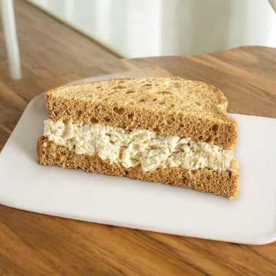 A Ranch Chicken Salad Sandwich on multigrain bread from E.A. Sween is placed on a white plate on a wooden table.