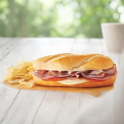 An Italian Style Sub by E.A. Sween, featuring sliced ham, cheese, and salami on a white baguette, served with a side of potato chips, placed on a wooden table with a blurred background.