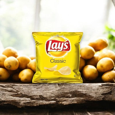 A bag of Lay's Classic Potato Chips 1 oz by Frito Lay sits in front of a pile of whole potatoes on a wooden surface with a blurred background, offering the perfect portion size for enjoying this beloved snack.