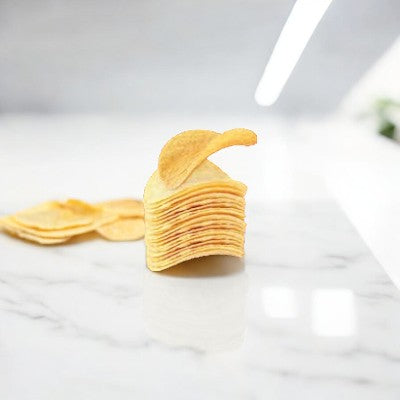 A stack of The Good Crisp Company Gluten Free Original Snack Chips stands on a white marble surface with some loose chips in the background. A brightly lit environment is visible.