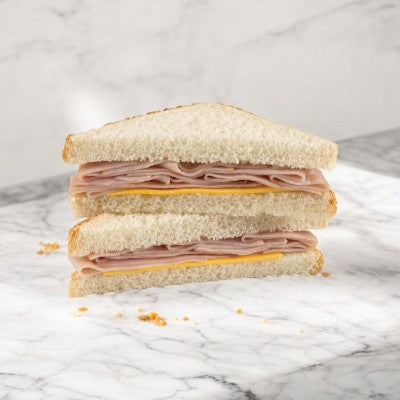 A Smoked Ham & Cheese Sandwich Wedge from E.A. Sween, cut into two triangles, each with layers of smoked ham and cheese nestled between slices of white bread, resting on a marble surface surrounded by some crumbs.