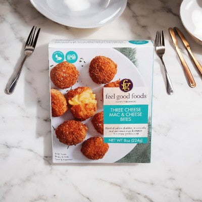 A box of "Easy Lunches Feel Good Foods Cheese Mac & Cheese" on a marble surface next to a fork, knife, and empty plate. The box shows an image of six creamy mac and cheese bites, the perfect comfort food for any occasion.