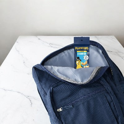 A packet from the Planters Cashews 5-pack (each bag 1.5 oz) is partially visible inside the open main compartment of a blue backpack placed on a white marble surface, showcasing Mr. Peanut’s quality snacks by Planters.