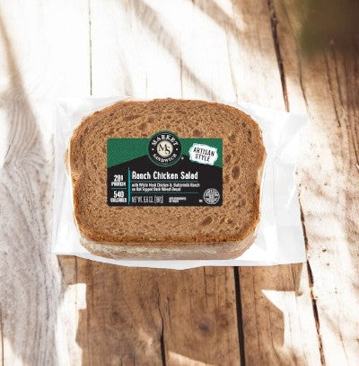 A packaged "Ranch Chicken Salad Sandwich On Multigrain Bread" from E.A. Sween on a wooden surface. The packaging displays nutritional information and "Artisan Style" branding.