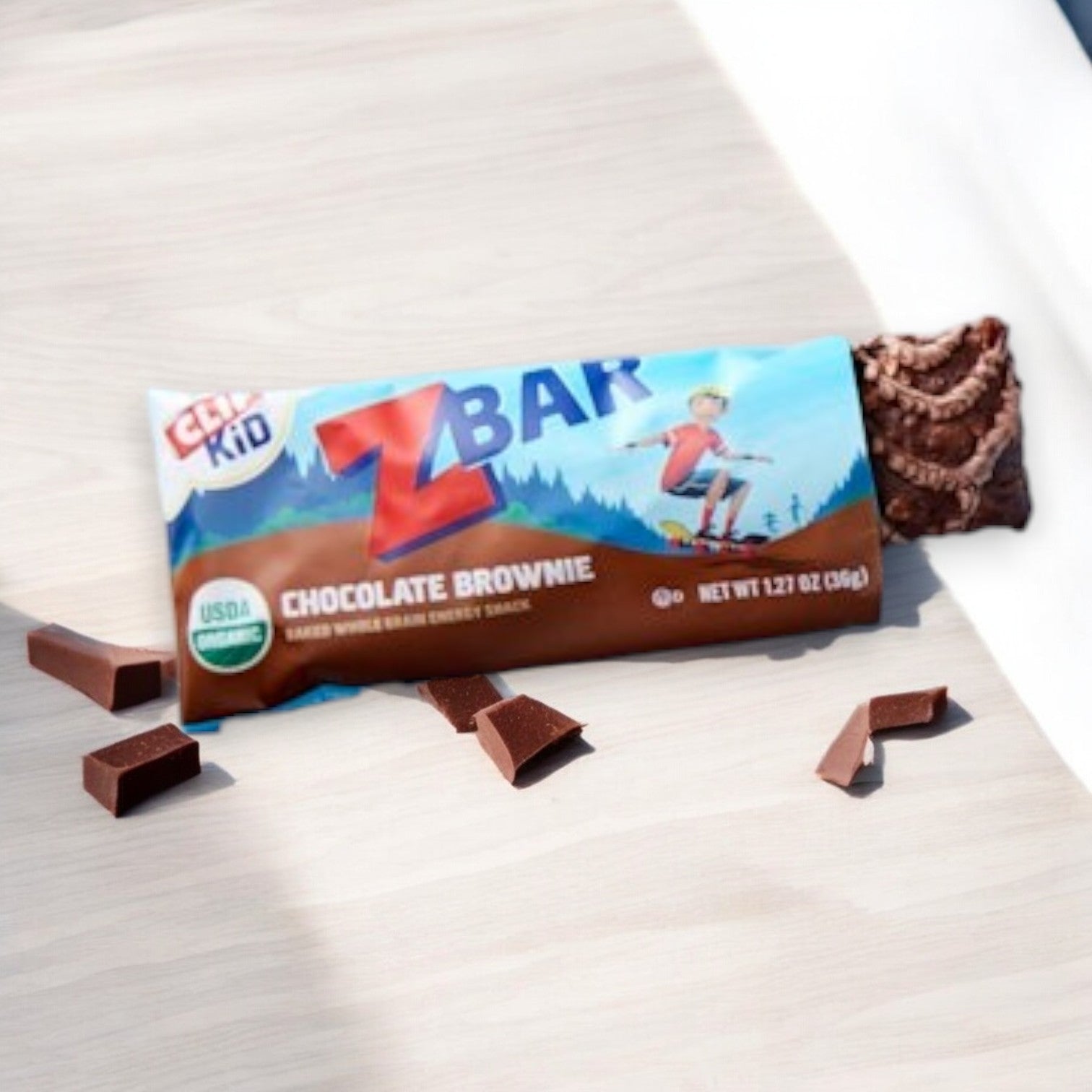 An open Clif Zbar - Chocolate Brownie, Soft Baked Whole Grain Snack Bar by Clif Bar (1.27 oz), sits invitingly on a light wooden surface with pieces of chocolate next to it, showcasing a nutritious snack bar perfect for kids.