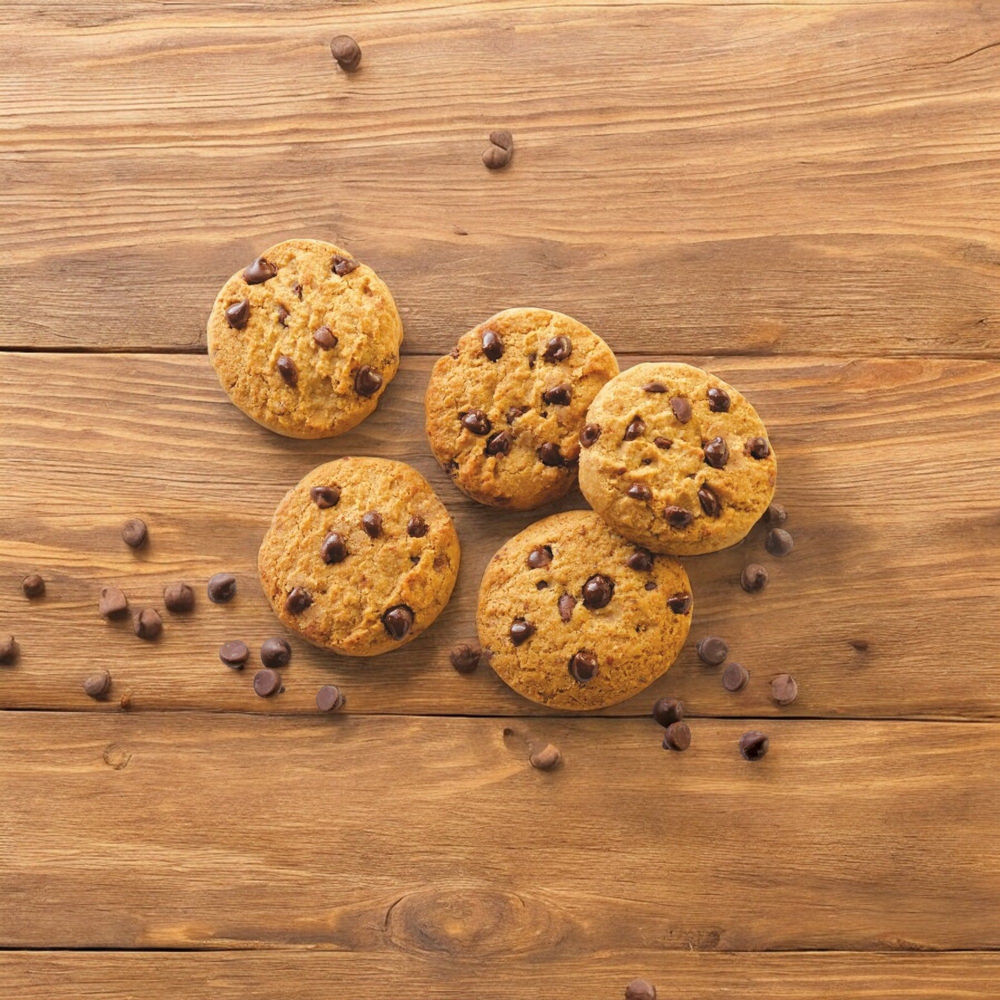 Five Simple Mills Chocolate Chip Cookies from a 5.5 oz box arranged on a wooden surface, with some chocolate chips scattered around.