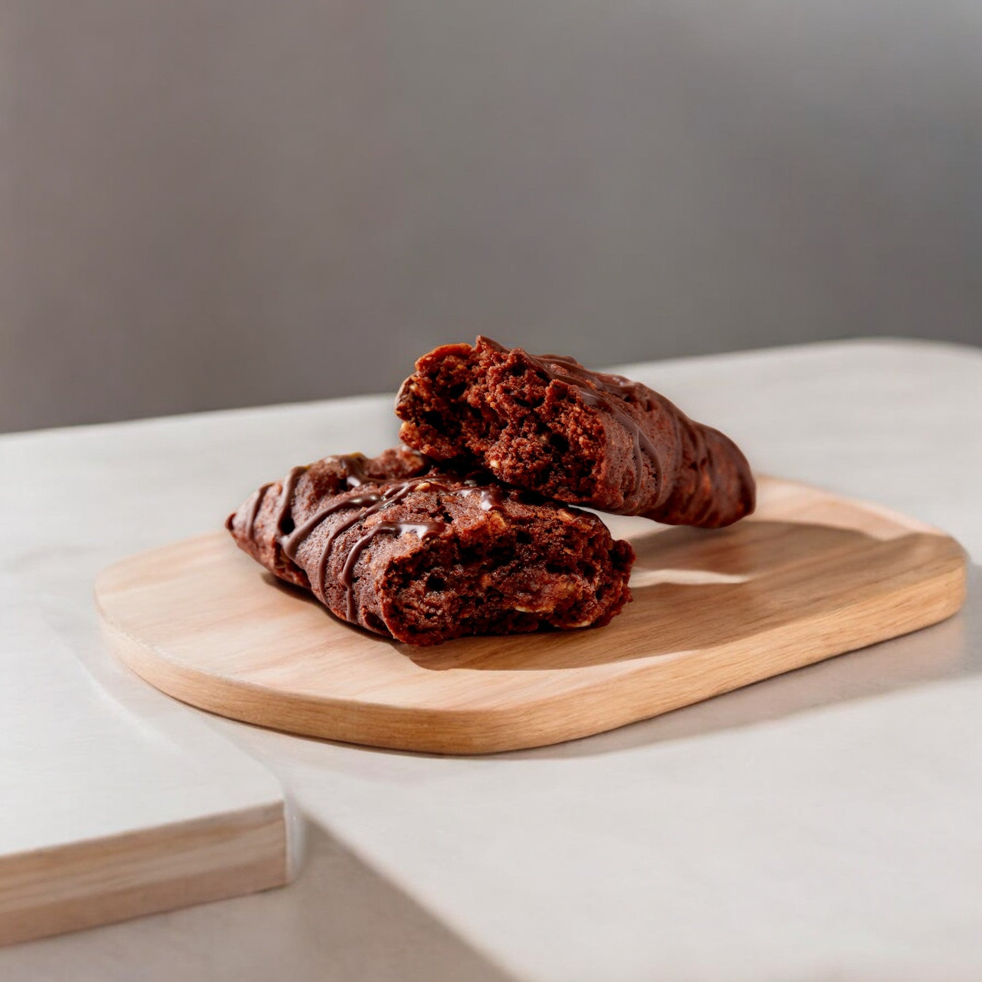 Two chocolate croissants are placed on a wooden board, with one croissant split open, revealing its rich, chocolate-filled interior—an indulgence that contrasts with the wholesome goodness of Clif Bar's Clif Zbar in Chocolate Brownie flavor, a soft baked whole grain snack bar weighing 1.27 oz.