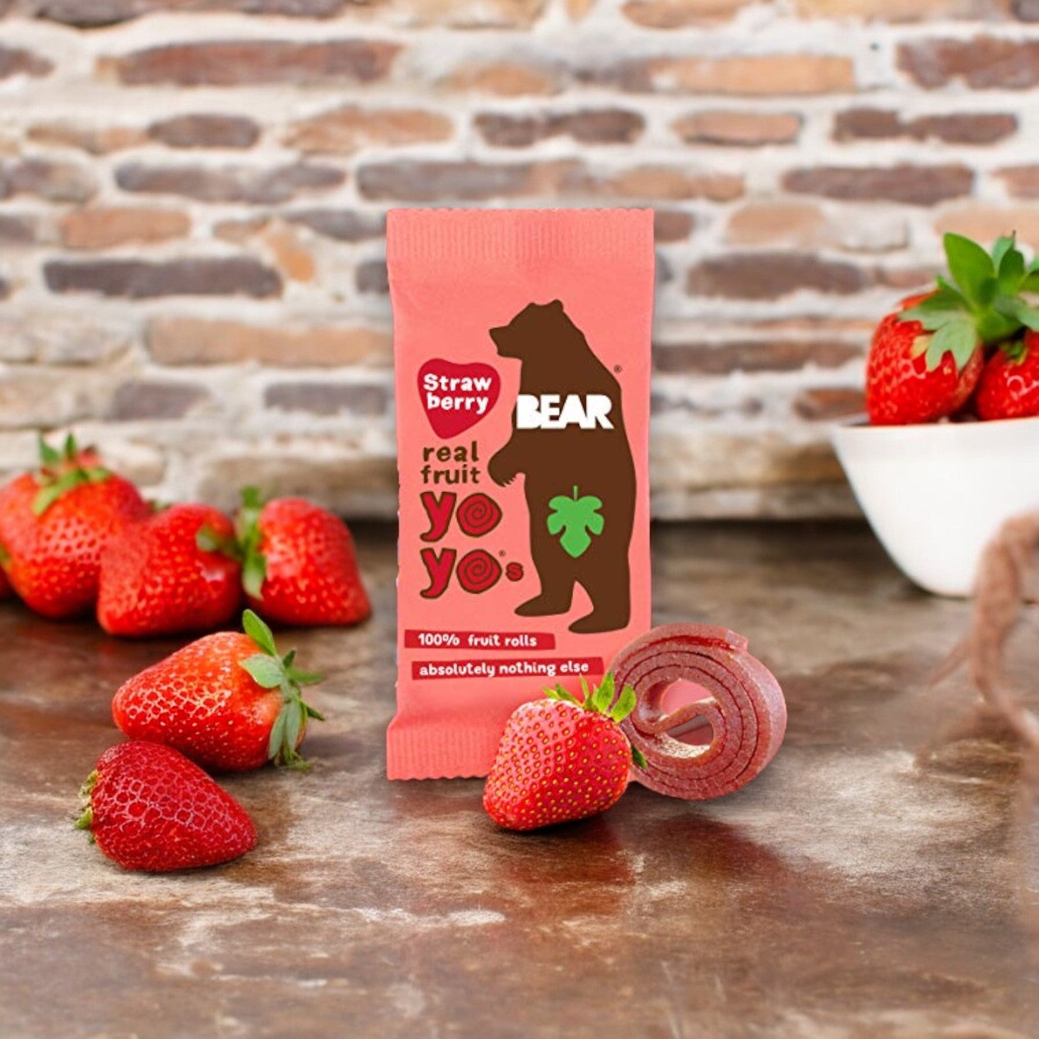 A package of Bear Fruit Rolls - Strawberry, 0.7 oz- 1 Count is surrounded by fresh strawberries and a small white bowl of strawberries in front of a brick wall, illustrating these natural fruit snacks as delicious and healthy options.