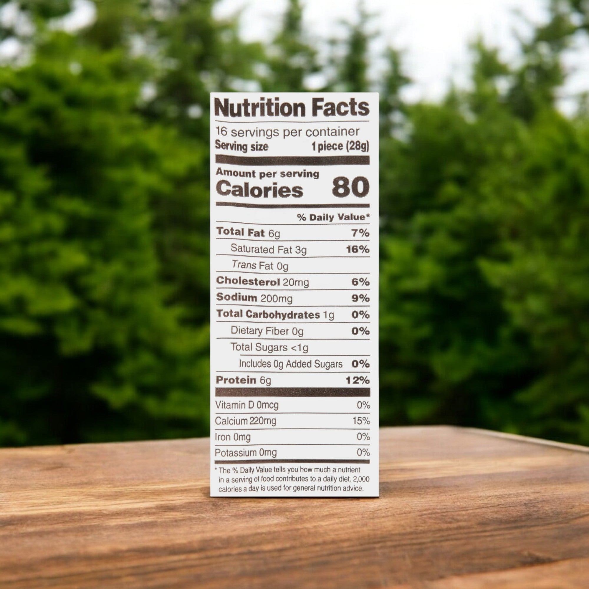 Close-up of a nutrition facts label on Frigo® Cheese Heads® Original Mozzarella String Cheese Snacks - 1 oz - 1 Count, a high-protein snack showing amounts per serving: 80 calories, 6g total fat, 6g total carbs, and 6g protein. Background features blurred outdoor scenery.