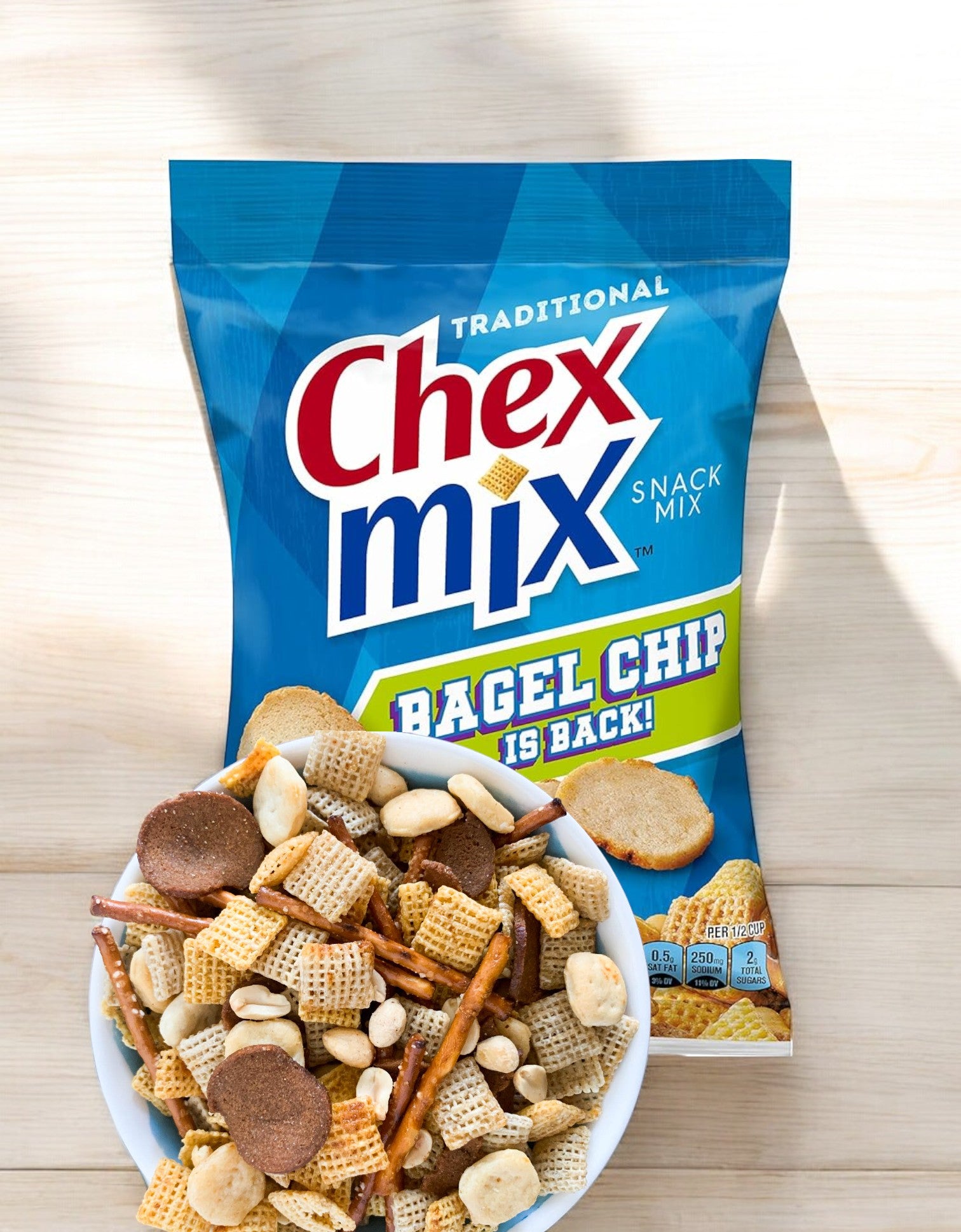 Behind a bowl brimming with assorted snacks like pretzels, bagel chips, and Chex cereal pieces, sits a bag of Chex Mix Traditional Savory Snack Mix by General Mills, featuring the announcement "Bagel Chip is Back!" Perfect for sharing, this Variety Pack offers 42 individual bags.