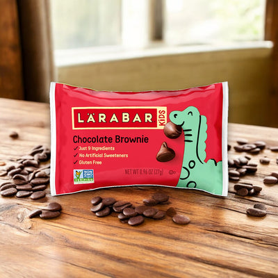 A package of Larabar Kids Chocolate Brownie Gluten Free Bars, 0.96 oz - 1 Count, rests on a wooden surface, with chocolate chips scattered around it. The packaging emphasizes being non-GMO, gluten-free, and containing no artificial sweeteners.