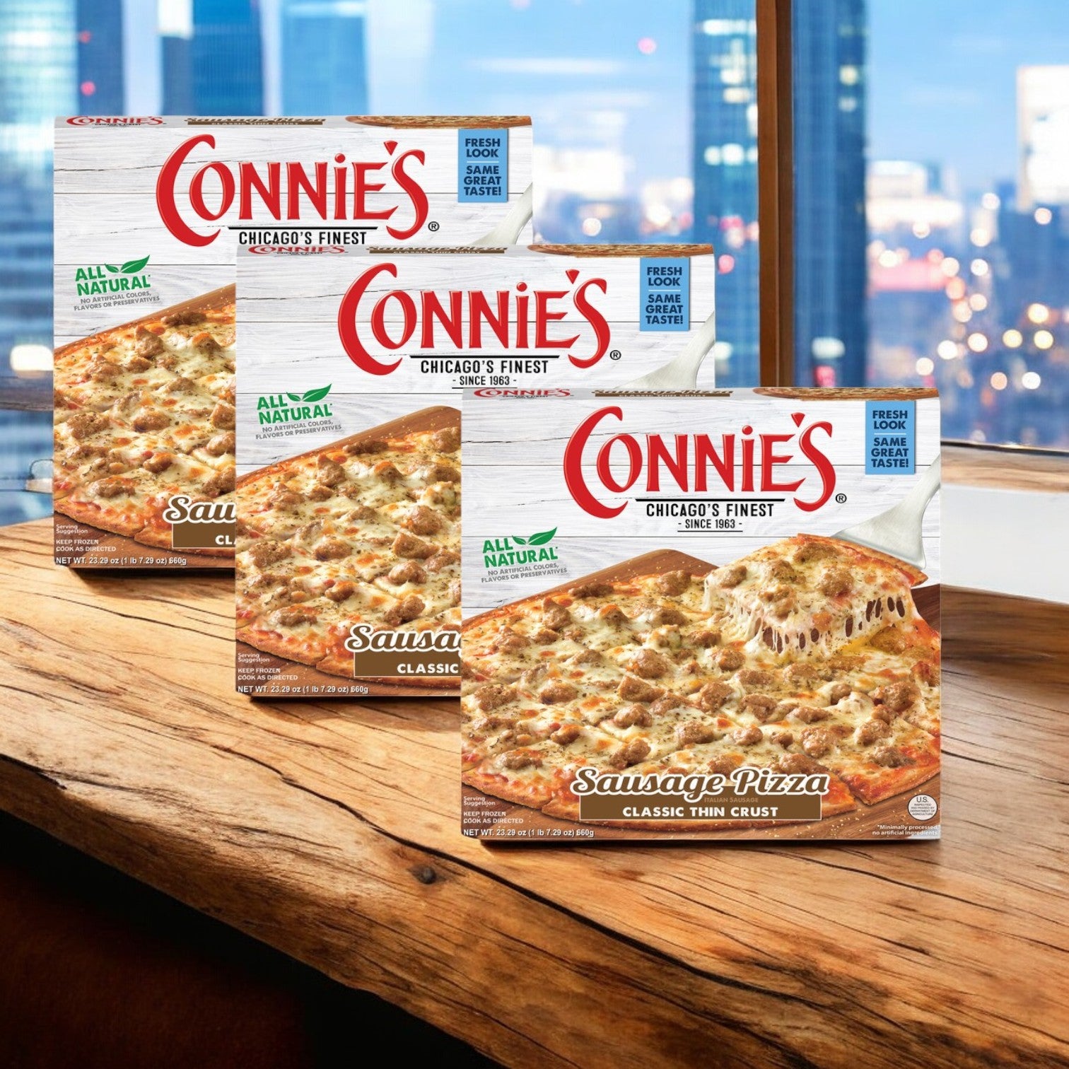 Displayed on a wooden table with a cityscape in the background are three boxes of Connie's Pizza Sausage Classic Thin Crust, 23.29 oz packs. The easy preparation of this delicious meal ensures you can enjoy it effortlessly.