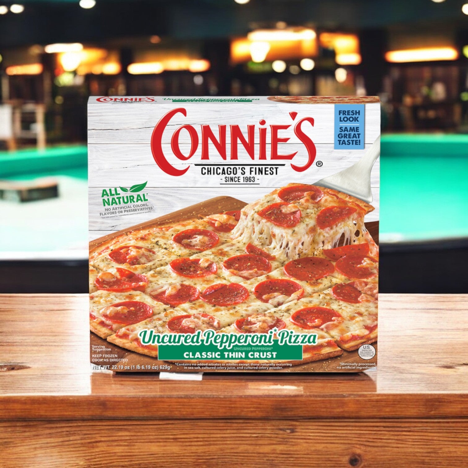 A box of Connie's Pizza Classic Thin Crust Uncured Pepperoni sits on a wooden table with a blurred pool table in the background, promising easy preparation and showcasing a delightful blend of savory sausage.